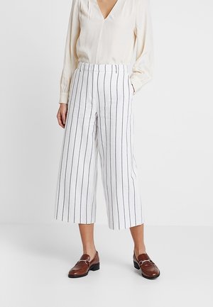 Frau trägt weiße, weite, verkürzte Hose mit schwarzen vertikalen Streifen, cremefarbene Bluse mit langen Ärmeln und braune Leder-Loafer.