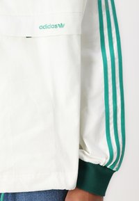 Biela bunda s zeleným logom Adidas, tromi zelenými pruhmi na rukávoch, ribbovanými manžetami a hladkým povrchom.
