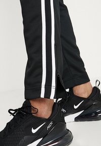 Svarta idrottsbyxor med vita sidosträck och blixtlås vid anklarna, tillsammans med svarta Nike Air Max 270 sneakers med vita swoosh-logotyper.