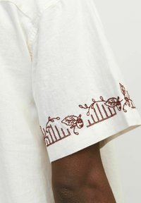 Hvid linnedskjorte med ærmer, der har intrikate broderier af kastanjer og egeblade i maroon, hvilket tilføjer tekstur og dekorative detaljer.