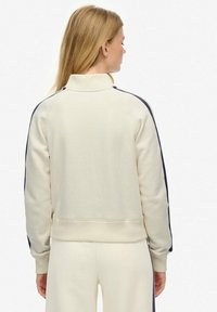 Sweatshirt crème avec un col haut, présentant des rayures latérales bleu marine et des poignets et un ourlet côtelés. Texture lisse, coupe décontractée.