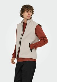 Beige ärmellose Weste mit Stehkragen, Frontreißverschluss, Seitentaschen und schwarzer Bekkung, getragen über einem rostfarbenen Langarmhemd.
