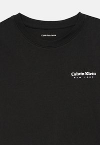 Černé bavlněné tričko s kulatým výstřihem, se symbolem bílého loga Calvin Klein New York na levé části hrudi. Hladký povrch.