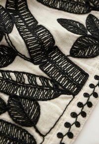 Feuilles noires brodées avec des coutures détaillées sur un fond en tissu crème, avec une bordure décorative de pois noirs et de boucles.