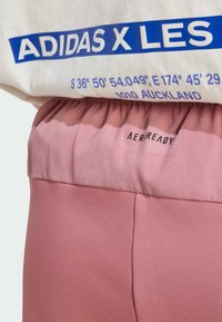 Das Adidas X Les T-Shirt besteht aus einem weißen Stoff mit blauem Grafikdruck. Darunter befinden sich rosa AEROREADY-Shorts mit einem gerafften Bund.