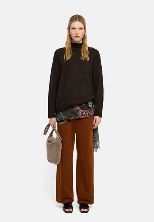 Kobieta w czarnym swetrze, z wzorzystym szalikiem, brązowych spodniach z szerokimi nogawkami, czarnych butach z odkrytymi palcami, trzymająca beżową zamszową torebkę na pasku.