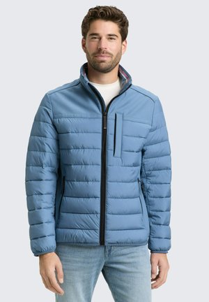 Mężczyzna w jasnoniebieskiej pikowanej kurtce z czarnym zamkiem błyskawicznym oraz zamykanymi kieszeniami na piersi i po bokach, stojący na tle jednolitego jasnego tła.