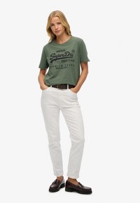 Grünes Baumwoll-T-Shirt mit schwarzer Vintage-Grafik; weiße Slim-Fit-Hose; brauner Ledergürtel; schwarze Slipper mit dicker Sohle.