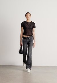 Čierny pruhovaný sieťovaný crop top, tmavé džínsy s vysokým pásom a rozšírenými nohavicami, biele tenisky a malá čierna kabelka, stojaca na svetlom povrchu.