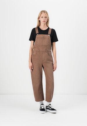 Brown overalls made of denim, featuring a front pocket, adjustable straps, and a relaxed fit, paired with a black t-shirt and sneakers.