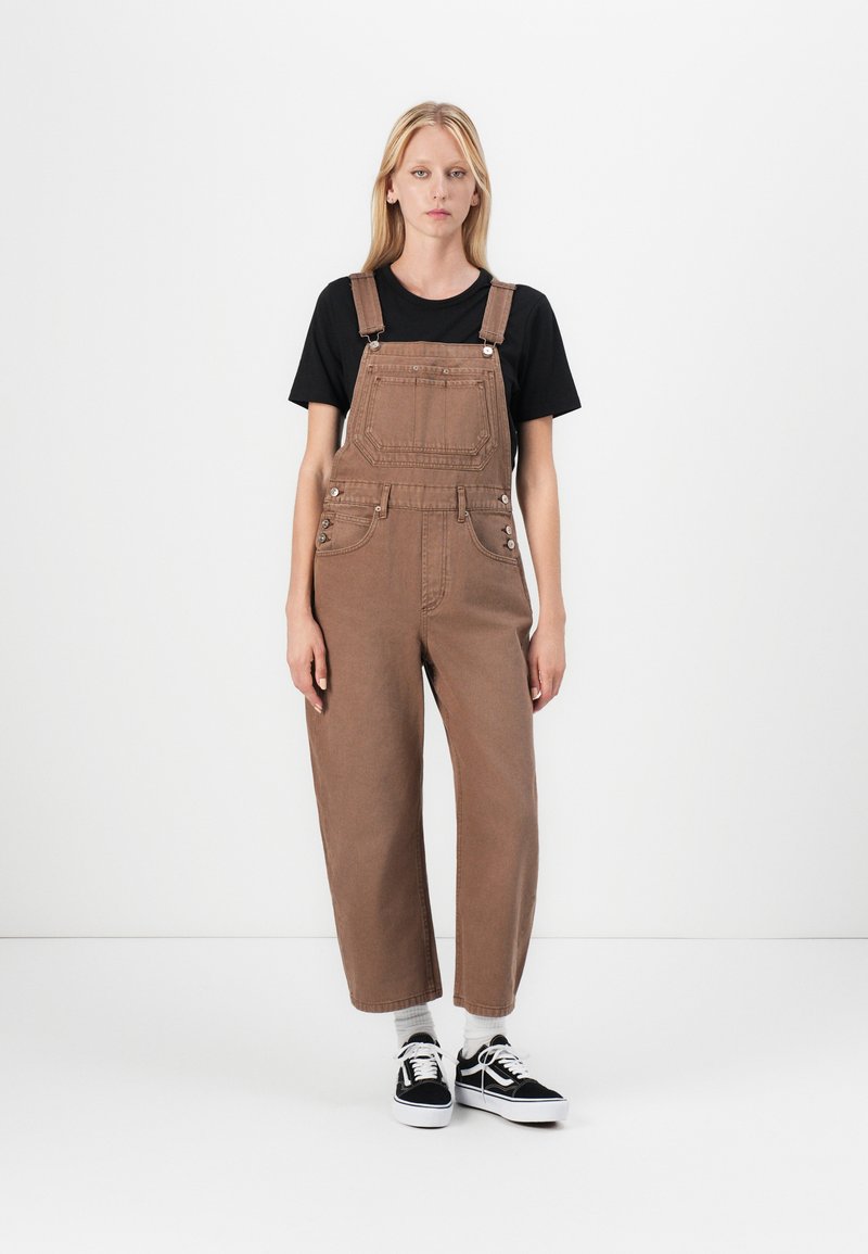 Bruine denim overall, voorzien van een voorzak, verstelbare bandjes en een ontspannen pasvorm, gecombineerd met een zwart T-shirt en sneakers.