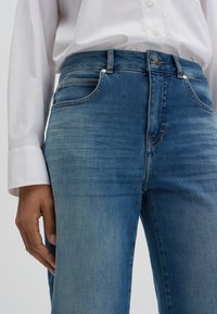 Jeans in denim blu con vita aderente, cinque tasche e leggere sfumature. Abbinati a una camicia bianca con bottoni, caratterizzata da una texture liscia.