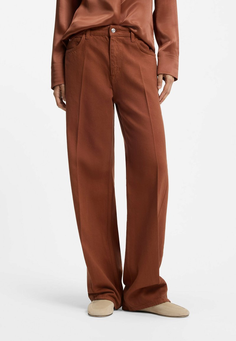 Personne portant un pantalon large marron ample, une chemise marron à manches longues assortie et des chaussures couleur crème, debout devant un fond uni.