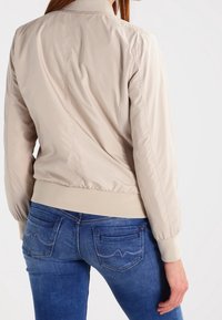 Ljusbeige bomberjacka med ribbade muddar och fåll, med en slät textur och elastiska sidopaneler. Matchad med blå jeans.