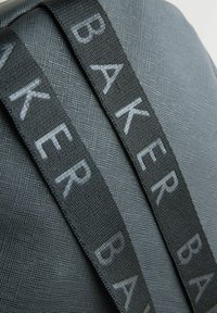 Černá taška s texturovanou tkání a výrazným šedým logem "BAKER" na nastavitelných popruzích. Elegantní obdélníkový tvar, minimalistický design.