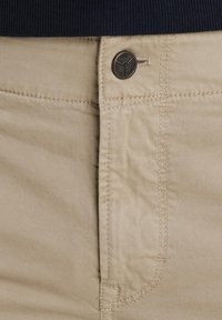Pantalon beige avec bouton visible et détails de couture, porté avec un haut à taille côtelée foncée.