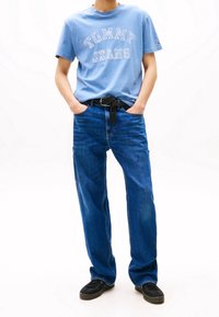 Camiseta de algodón de color azul claro con estampado blanco de "TOMMY JEANS". Jeans de mezclilla de color azul oscuro, de corte holgado, con bolsillos laterales. Cinturón negro y zapatillas negras.