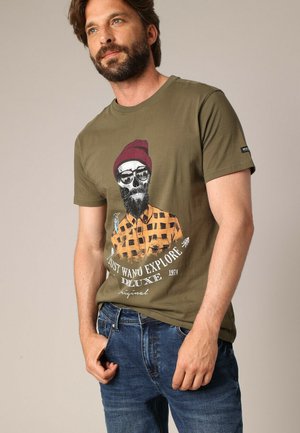 Homme portant un t-shirt vert olive avec un graphique de crâne, un bonnet rouge et une chemise à carreaux, associés à un jeans bleu, se tenant avec une main dans la poche.