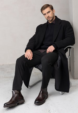 Brązowe, skórzane męskie botki sięgające kostki z suwakiem z boku, czarne spodnie i czarny płaszcz na ciemnym swetrze, siedzący na chromowanym krześle.