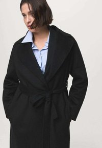 Manteau enveloppant noir en laine avec des revers larges, taille nouée et poches latérales, superposé sur une chemise rayée bleu clair. Texture douce, design élégant.