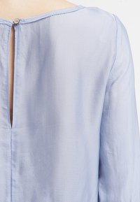 Ljusblå blus med en texturerad tyg och en nyckelhålsdetalj bak samt en enkelt knappstängning vid halslinningen. Långa, svävande ärmar.