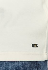 Katoenen T-shirt in gebroken wit met een gladde textuur, voorzien van een zwart label met gouden accenten nabij de zoom. De zoom is recht gestikt.