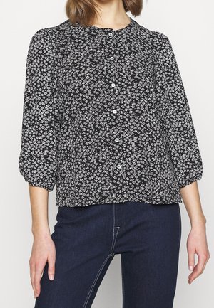 Kvinde iført en sort bluse med lange ærmer med lille hvidt blomsterprint og hvide knapper, kombineret med mørkeblå jeans.