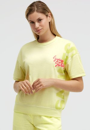 Young woman with straight blonde hair wearing a light yellow short-sleeve shirt and matching pants, hands gently clasped in front.