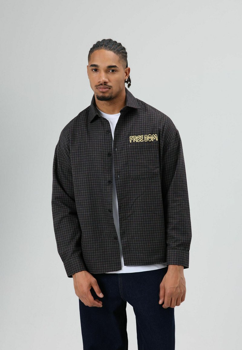 Geruit zwart overhemd met gele tekst "FREE DOM" op het linkerborstzakje, met een relaxte pasvorm en knoopsluiting aan de voorkant, gedragen over een wit shirt.