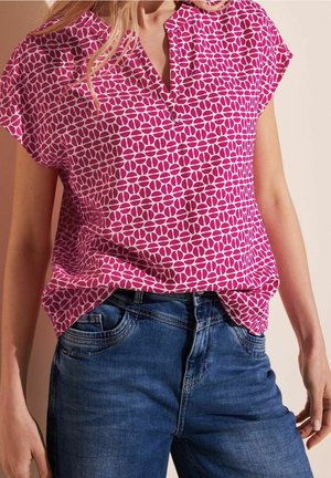 Femme portant un chemisier à manches courtes rose avec un motif géométrique de feuilles, rentré dans un jean en denim bleu, debout devant un fond neutre.