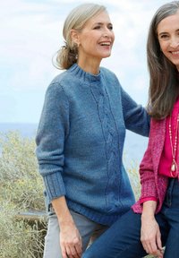 Blauer, gestrickter Pullover mit Zopfmuster, gerippten Bündchen und runder Ausschnitt; zeichnet sich durch ein weiches, strukturiertes Aussehen aus. Getragen mit grauen Hosen.