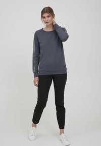 Felpa navy con maniche lunghe, caratterizzata da strisce bianche lungo le braccia e polsini a costine, abbinata a pantaloni neri e sneakers bianche.