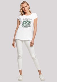 Hvit bomulls t-skjorte med et grønt grafisk trykk som har teksten "Malfoy 07 Slytherin." Kombinert med hvite slim-fit bukser og hvite joggesko.
