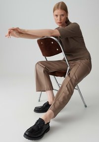 Bruin geribde gebreide top, beige synthetische broek en zwarte schoenen met franjes. Model zit op een metalen vouwstoel met een corduroy zitting.