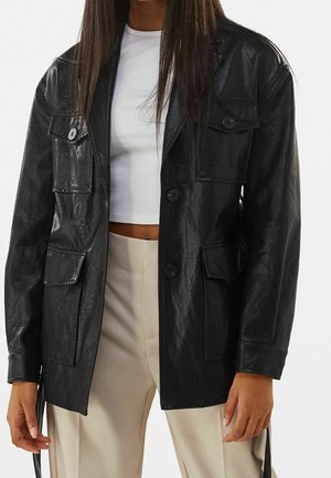Personne portant une veste en cuir noire par-dessus un crop top blanc et un pantalon beige taille haute, debout devant un fond uni.
