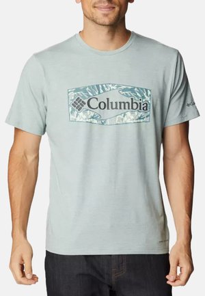 Mann trägt ein hellblaues Columbia-T-Shirt mit gemustertem Logo auf der Brust und dunkle Hose, steht vor einem einfarbigen Hintergrund.