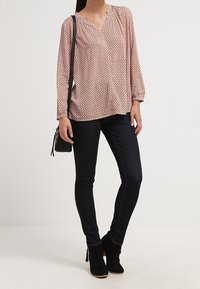 Blusa a maniche lunghe, scollo a V, di colore rosa chiaro con un motivo geometrico; abbinata a jeans skinny scuri e stivaletti neri, mentre tiene una borsa piccola.