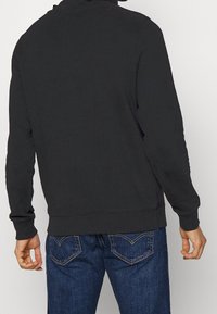 Felpa nera con colletto alto, maniche lunghe e polsini a costine. Abbinata a jeans blu scuro con tasche posteriori cucite.