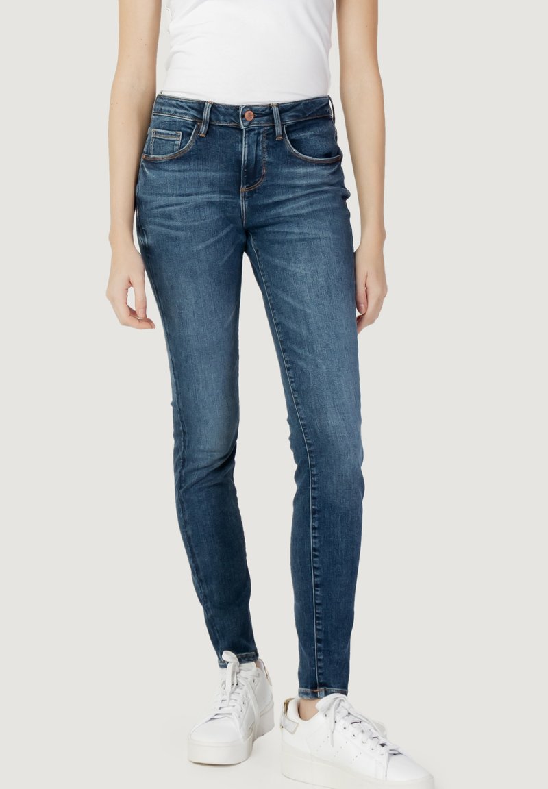 Tynne jeans i mørk blå denim med en glatt tekstur, fem lommer og subtil kontrastsøm, parret med hvite sneakers.