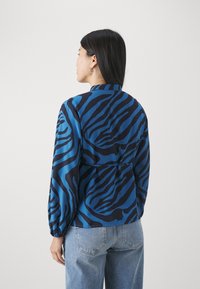 Blå og svart zebra-stripet bluse med lange ermer, med krage og rynket detalj på baksiden. Matchet med lyseblå jeans.