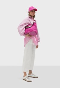Rosa übergroßer Pullover über einem durchsichtigen rosa Oberteil, weiße weit geschnittene Hose, weiße Sneakers mit Leopardenmuster und eine rosa Kappe. Rosa Handtasche präsentiert.