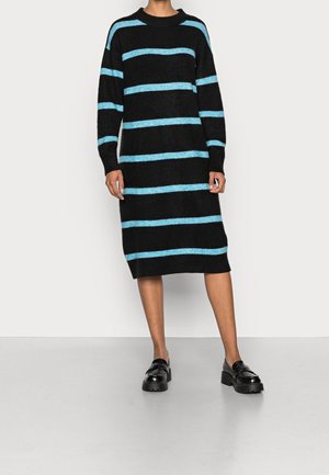 Schwarz gestreichtes Kleid mit horizontalen hellblauen Streifen, langen Ärmeln, Rundhalsausschnitt und knielanger Schnittform. Getragen mit schwarzen klobigen Schuhen.