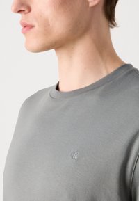 Cămașă T-shirt din bumbac gri, cu decolteu rotund, având un mic logo brodat pe pieptul stâng. Textură netedă și croială lejeră.