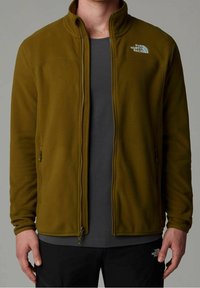 Man wearing olive green zip-up fleece jacket over dark gray shirt, with black pants, standing against plain gray background.