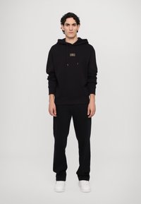 Black hoodie with a small gold logo, paired with black pants featuring white stitching and worn with white sneakers.