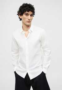 Άνδρας με σγουρά μαλλιά, φορώντας άσπρο πουκάμισο με κουμπιά και στρογγυλό λογότυπο στο στήθος και μαύρο παντελόνι, στέκεται με τα χέρια στις τσέπες.