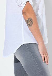 Hvid lærredsskjorte med optrukne ærmer, sideslidser, parret med lysegrå jeans. Bemærkelsesværdig tatovering synlig på armen. Nærbillede.