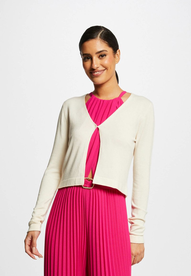 Cardigã creme com mangas longas, design aberto à frente e um único botão. Usado sobre um vestido rosa plissado com decote redondo e detalhes de alças.