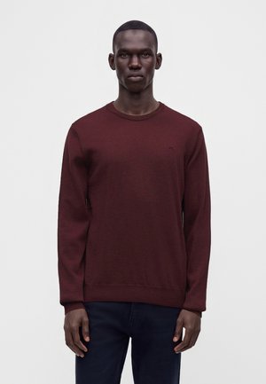 Homme portant un pull à manches longues uni rouge foncé et un pantalon sombre, debout devant un fond clair uni.