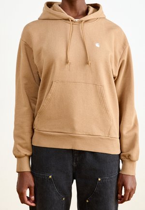 Homme portant un sweat à capuche beige avec poche kangourou et petit logo blanc, associé à un pantalon noir avec des poches rivetées.